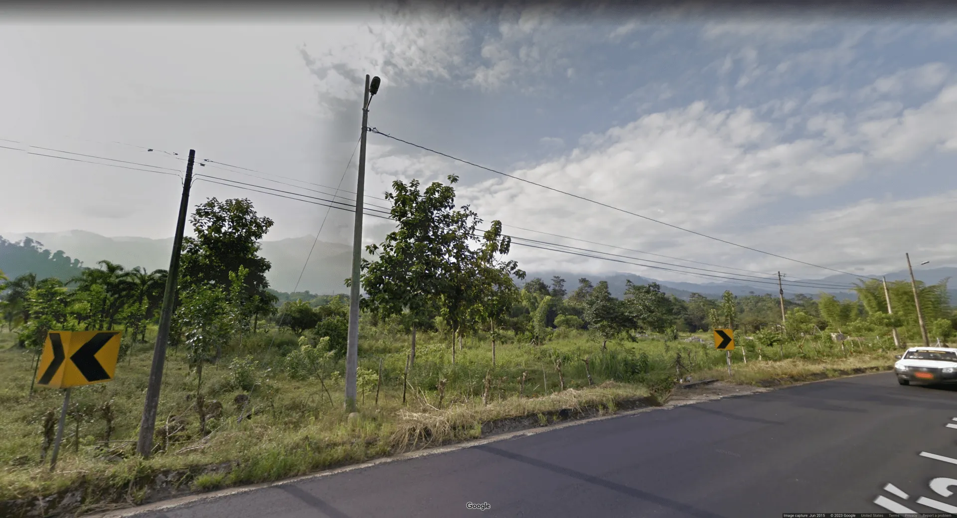 Picture of a roadside in a seemingly tropical area. There are black on yellow turn chevrons on the edges of the road, and a car off to the right with an orange license plate